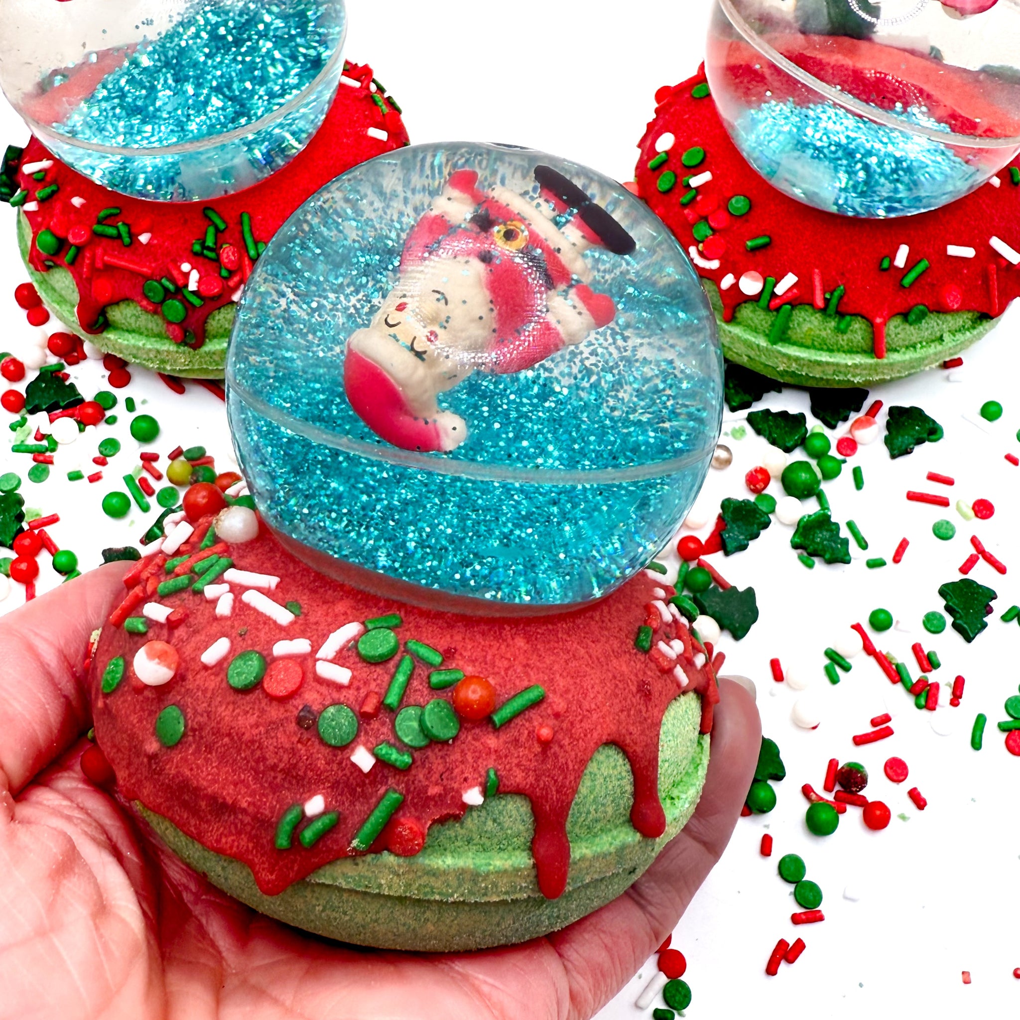 A Santa snow globe bath bomb in a hand. Green donut shaped bath bomb with red glaze topped with a Santa bouncy ball. Surrounded with red and green festive sprinkles.