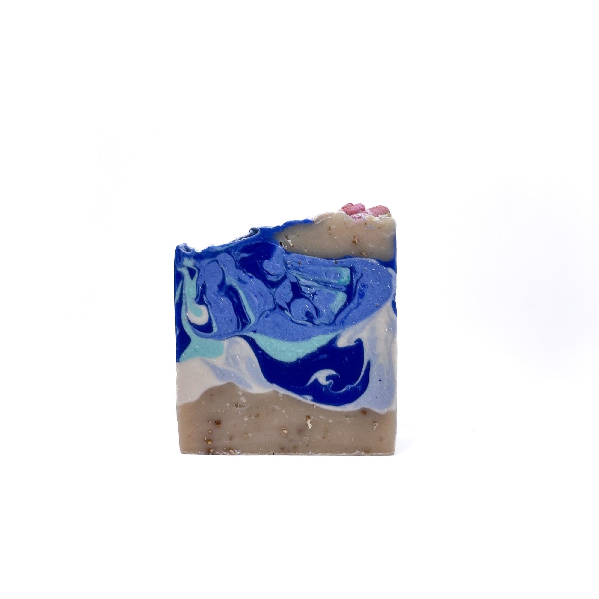 A bar of handmade soap. Tan with oatmeal grounds on the bottom for sand, Swirled white, teal, blue, and lite blue. Topped with sea shell soap embeds.