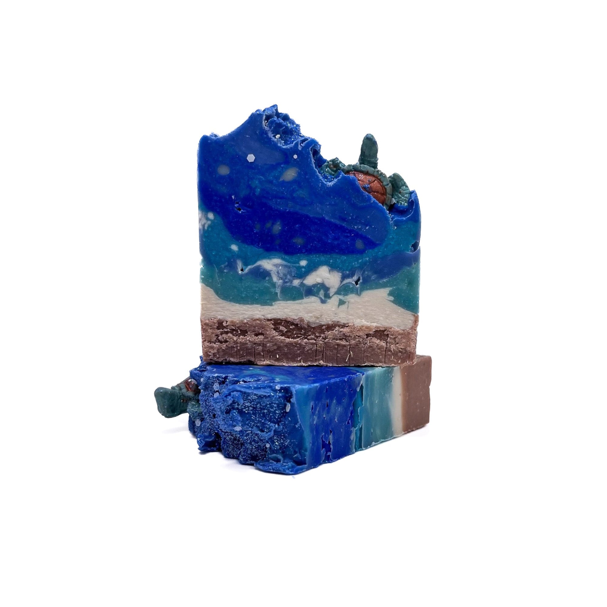 Two bars of Rusted Acre Soap Company's Sea Turtle handmade soap. One lying flat one upright. Tan bottom, swirled turquoise, blue and white. Topped with a sea turtle embed.