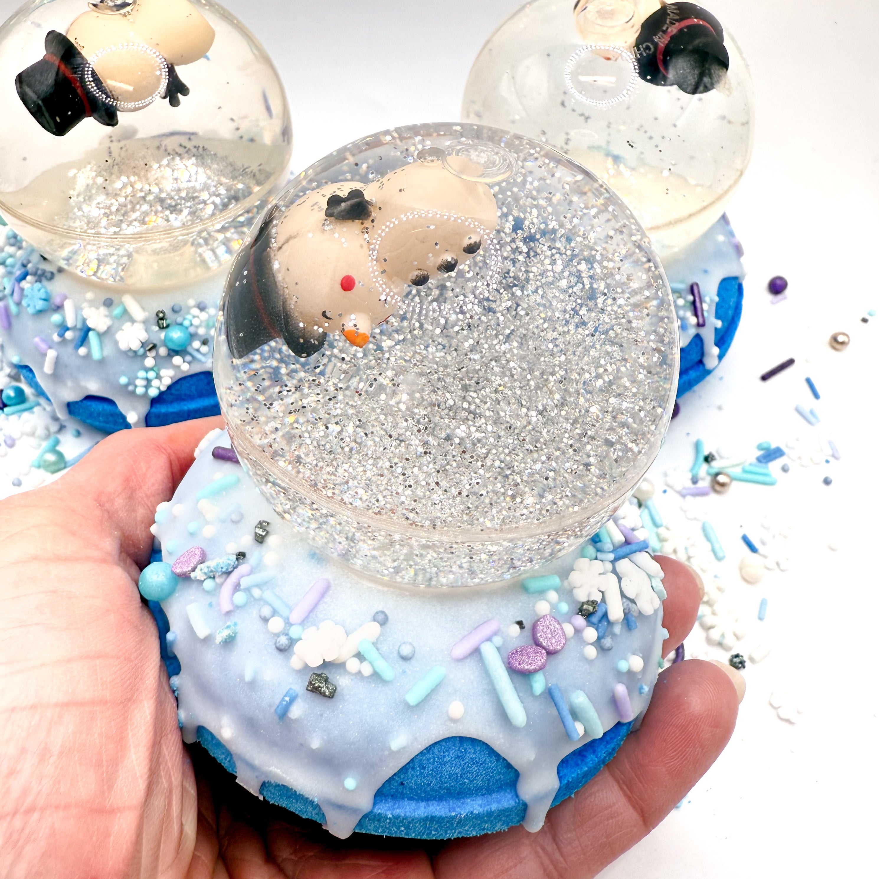 Snowman snow globe bath bomb being held in a hand. Bath bomb is a blue doughnut with white frosting and holiday sprinkles. Snowglobe is a bouncy ball with silver glitter photographed on a white background.