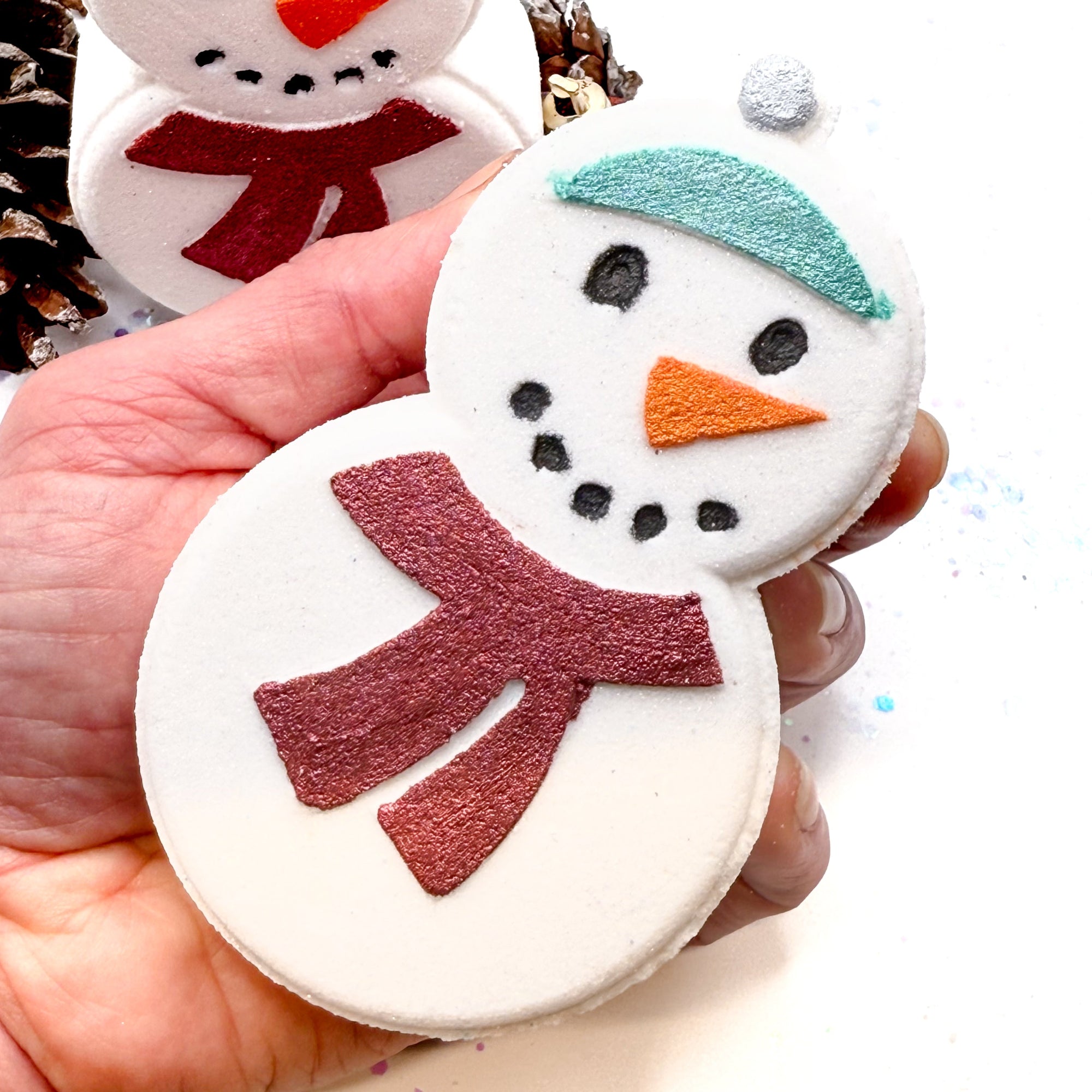 Hand holding a hand painted snowman shaped bath bomb made by Rusted Acre Soap Co with a red scarf and green hat.