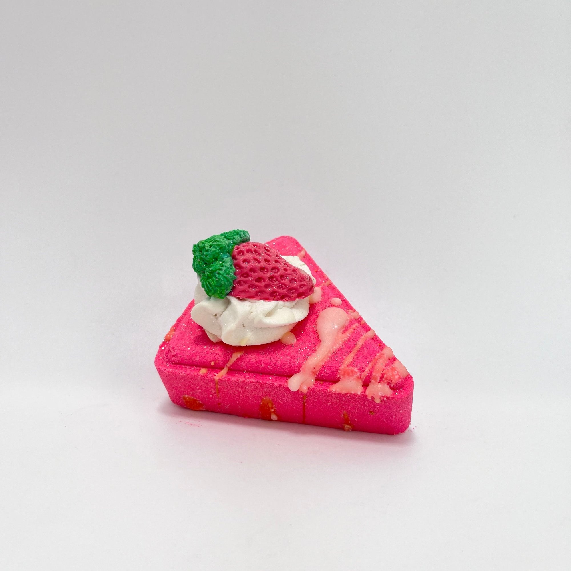 A pink strawberry pie slice shaped bath bomb. Topped with white bubble bar and a strawberry soap embed.