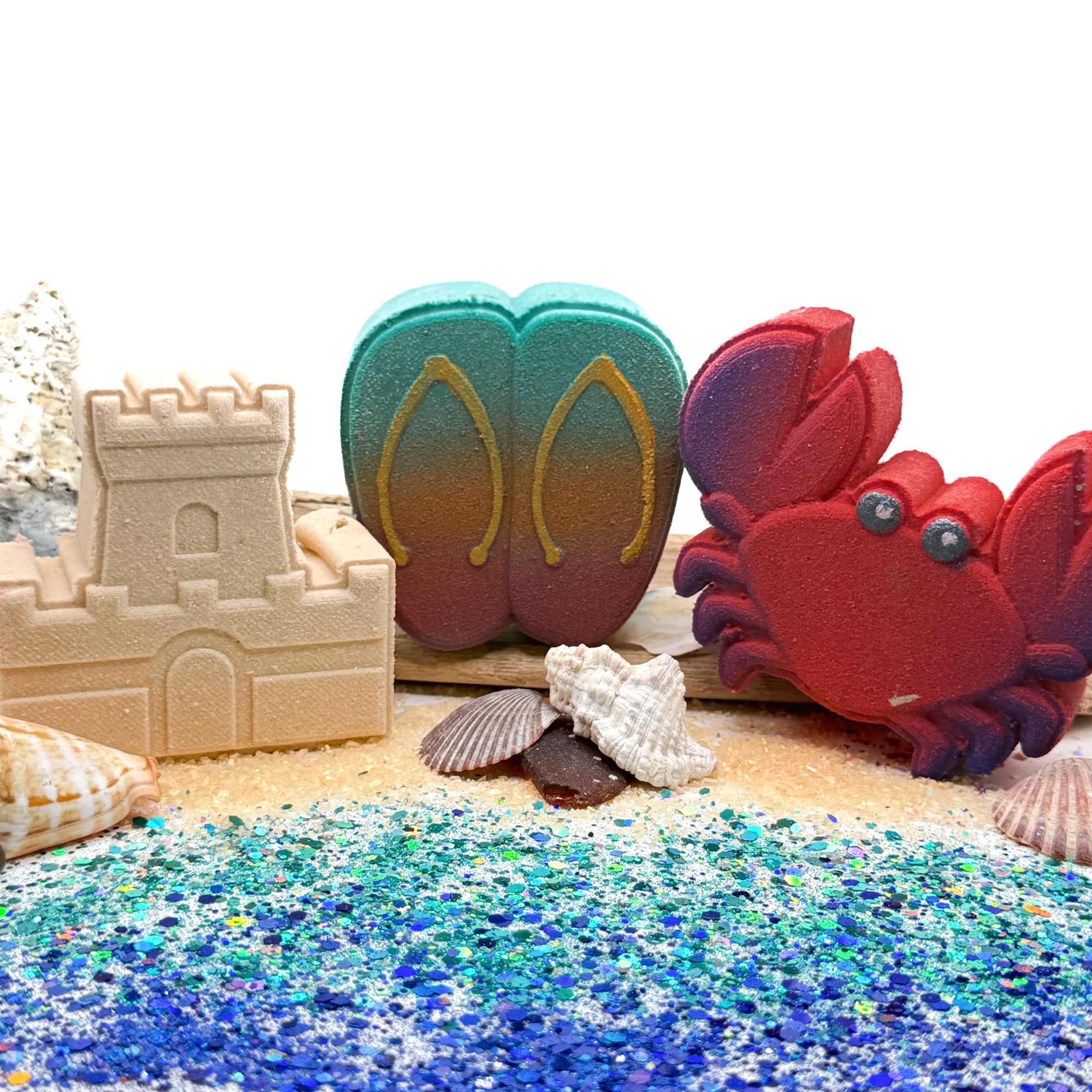 Three handmade summer themed bath bombs including flip-flops, a crab and a sandcastle on a sandy surface with shells and a sandcastle.