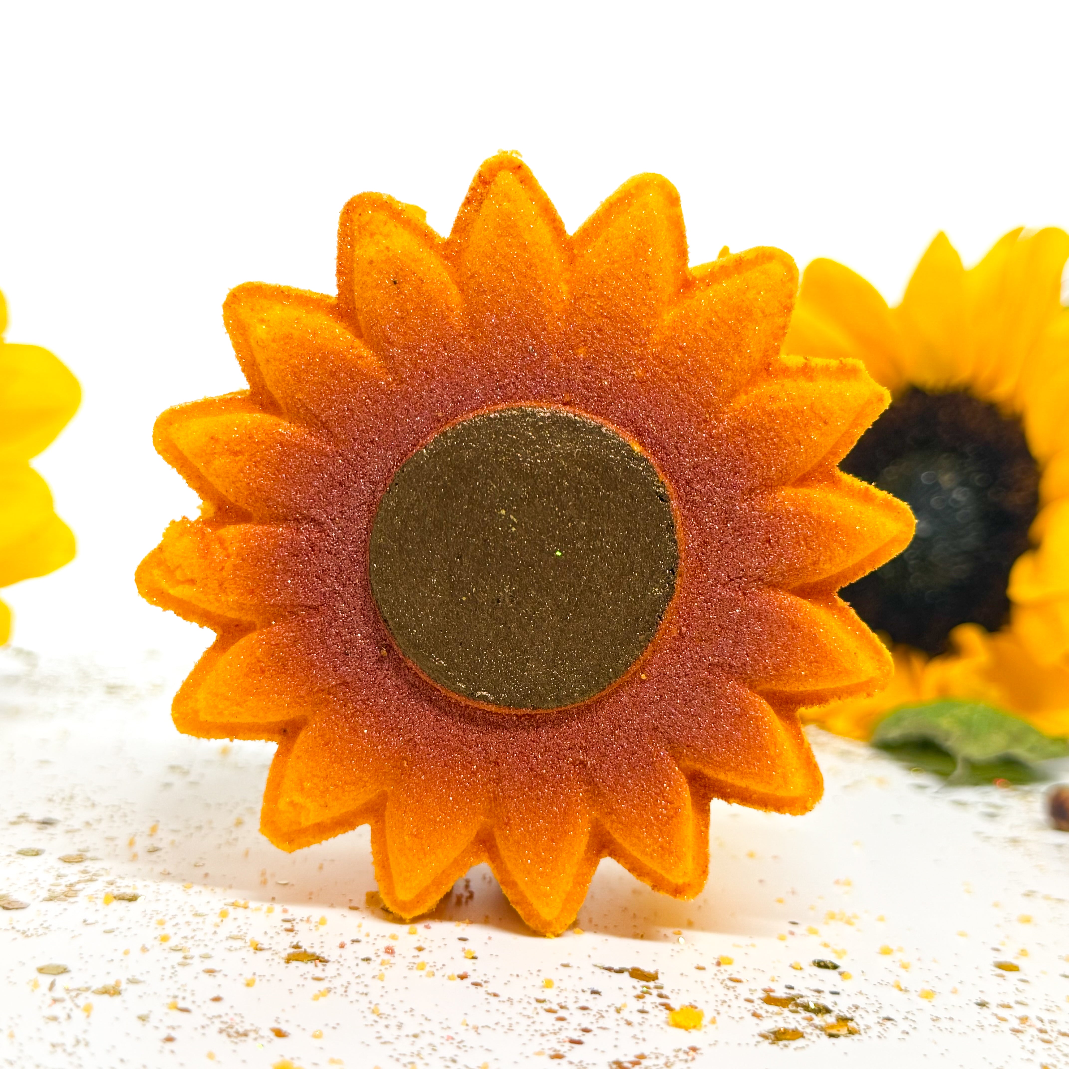 A close-up of a sunflower-shaped bath bomb with orange petals, a dark brown center, and a blurred second sunflower in the background on a white surface.