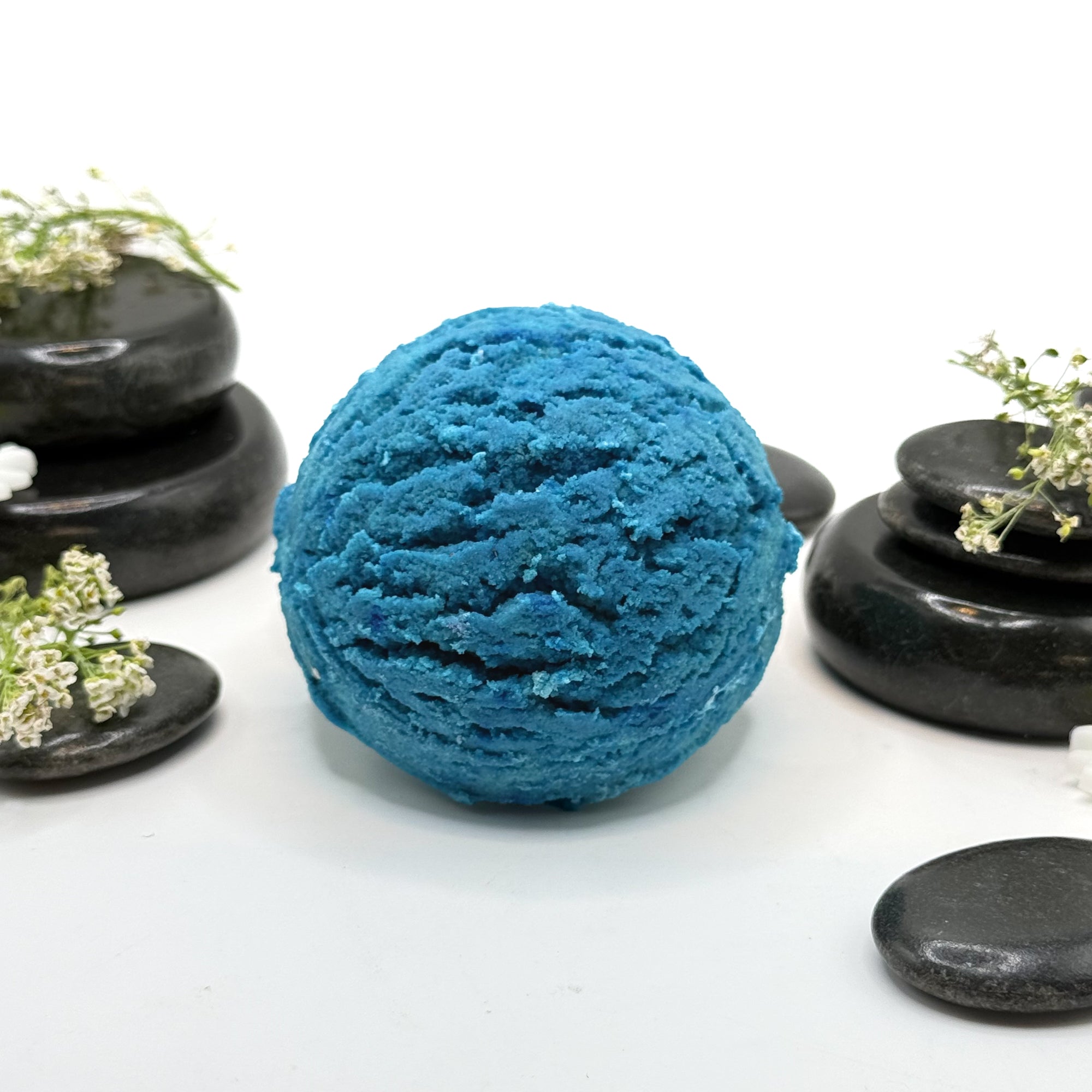 One blue bubble bath scoops that look like ice cream with spa stones and loose botanicals for a backdrop.
