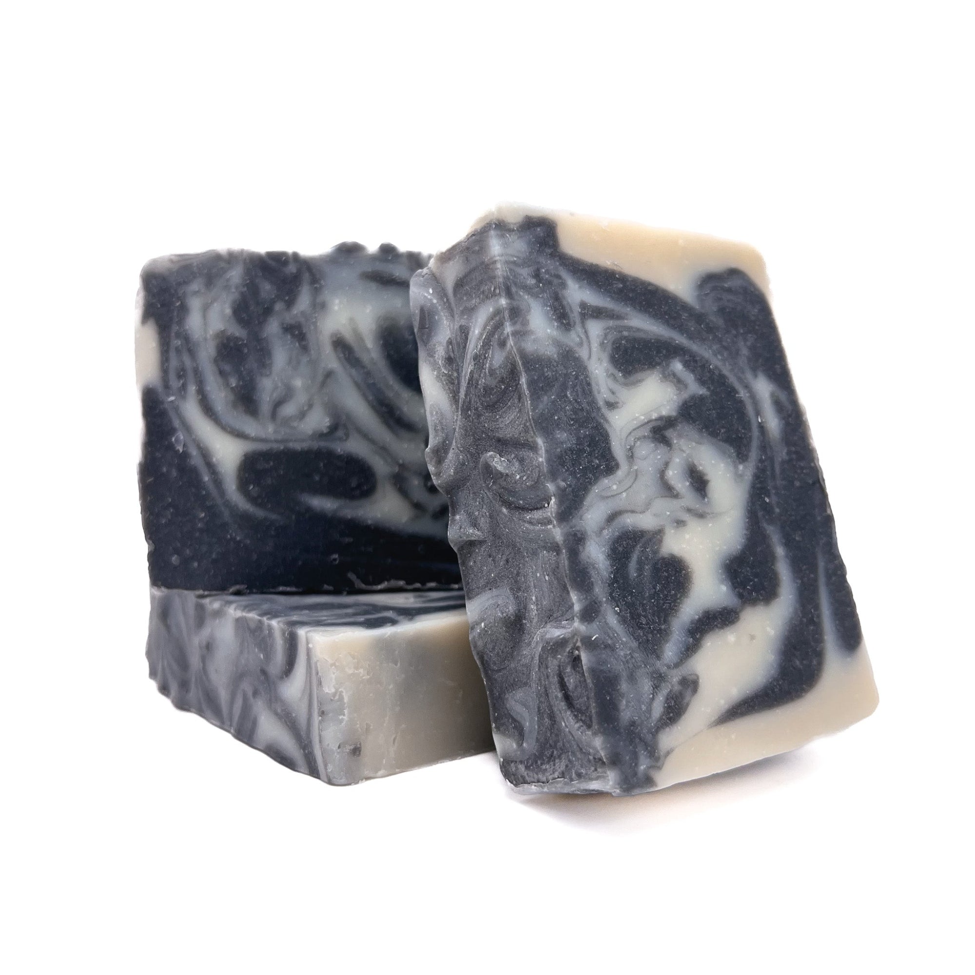 Three bars of Rusted Acre Soap Company's Tuxedo handmade soap. Charcoal black and natural white swirled. One lying flat two upright to show texture.