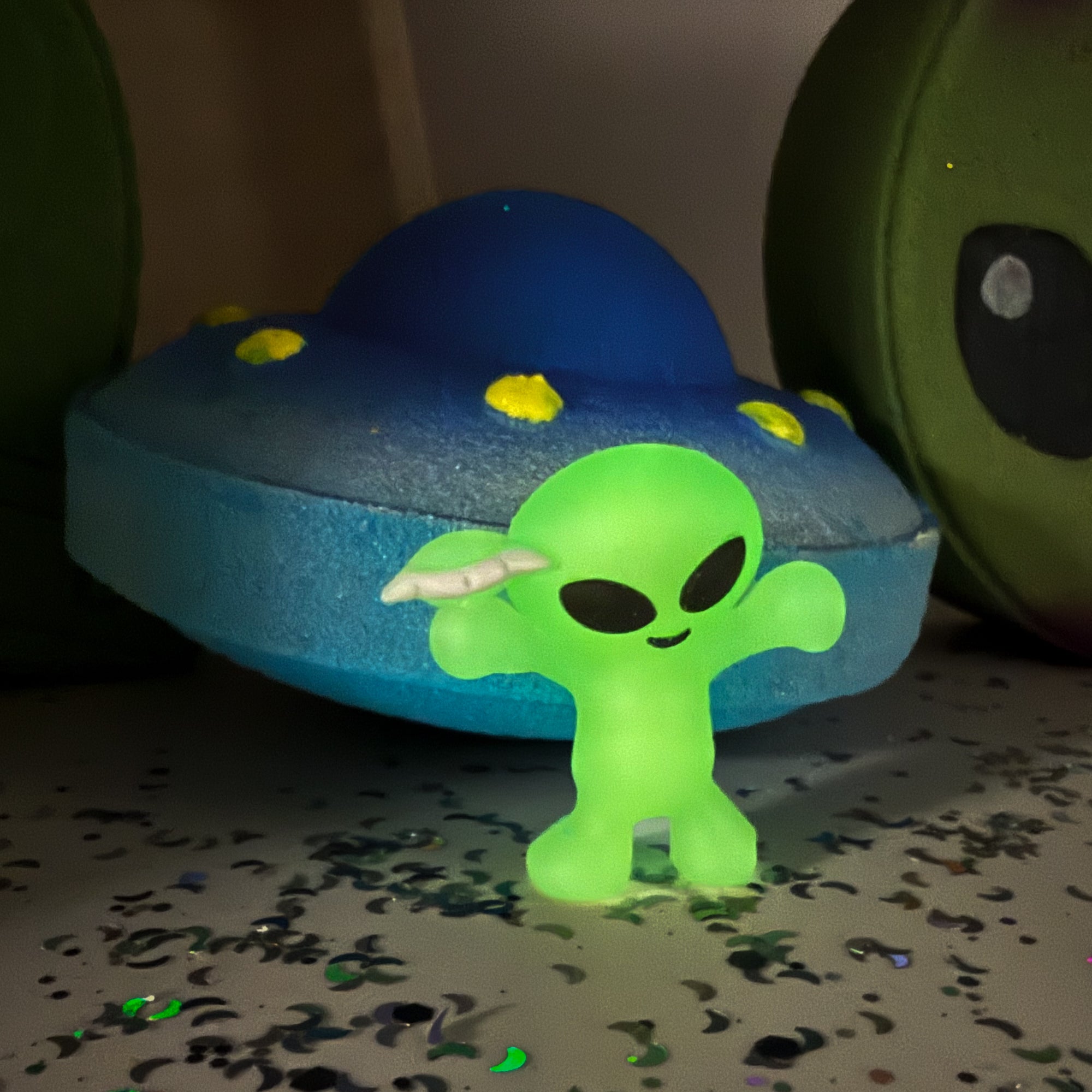 Glowing green alien figure with a blue UFO bath bomb in the background on a dark surface.