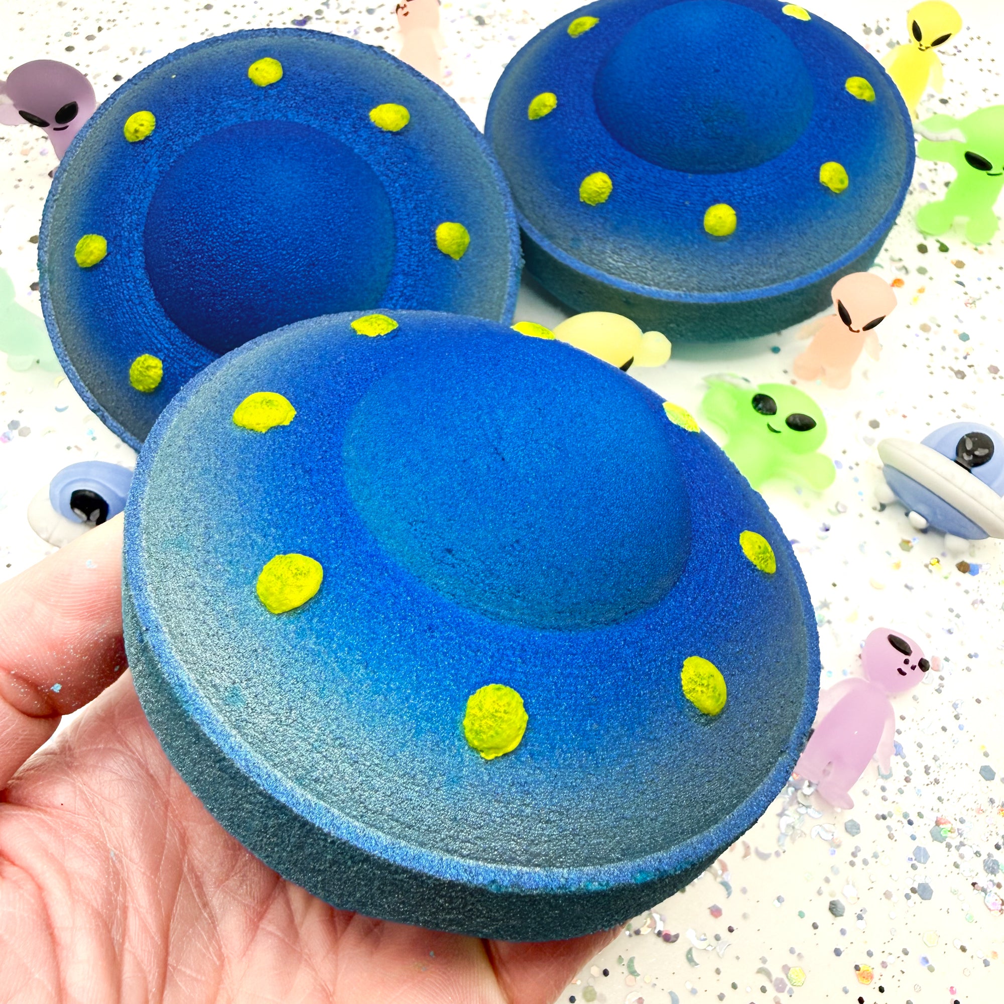 Blue UFO shaped bath bomb with yellow dots held in a hand against a colorful background.
