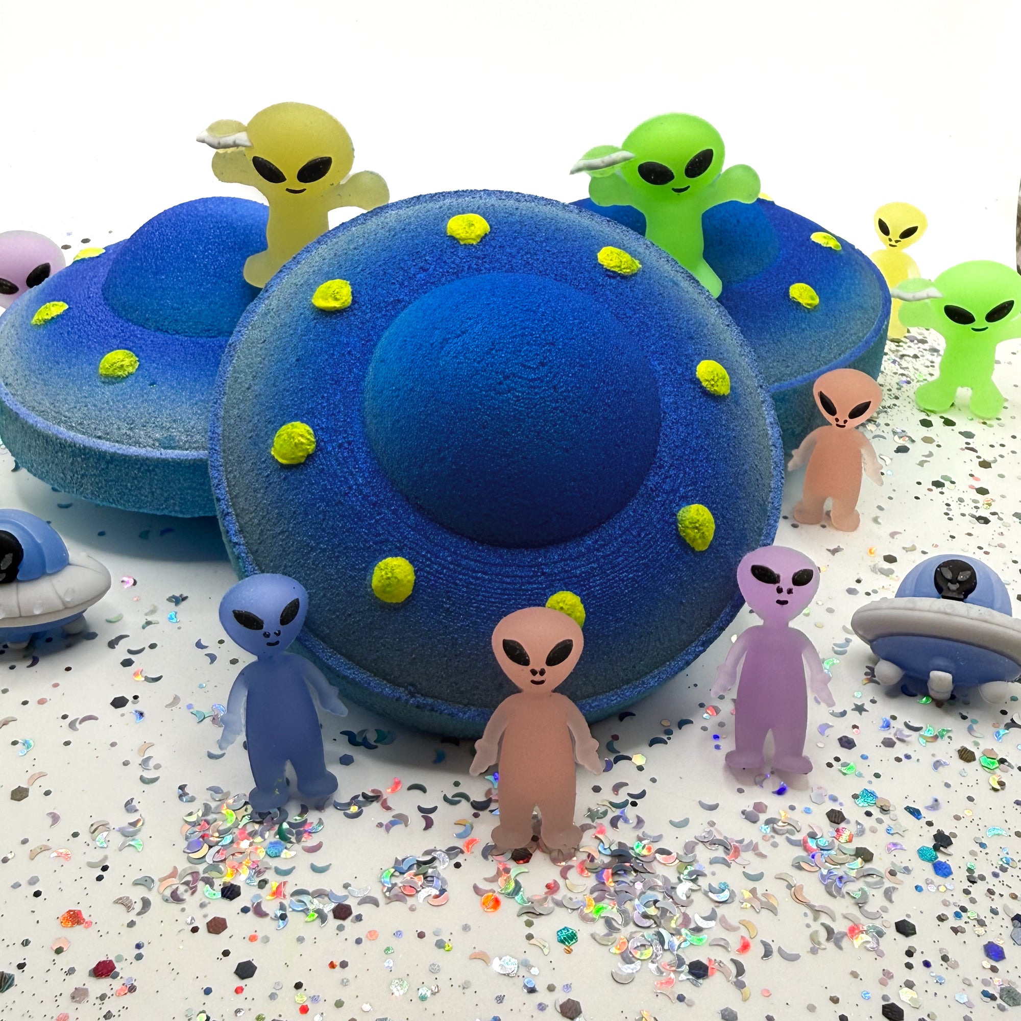 One blue and yellow UFO bath bomb showing the front profile with two bath bombs on a white background with alien toys placed around the bath bombs.