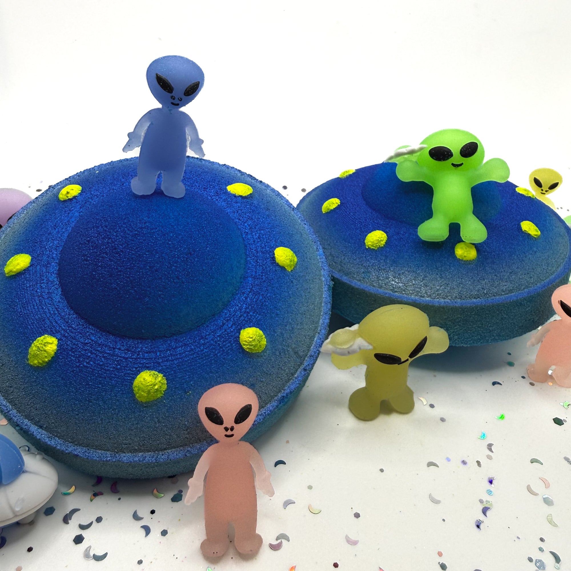 Two blue and yellow UFO bath bombs showing the top and side view on a white background with alien toys placed around the bath bombs.