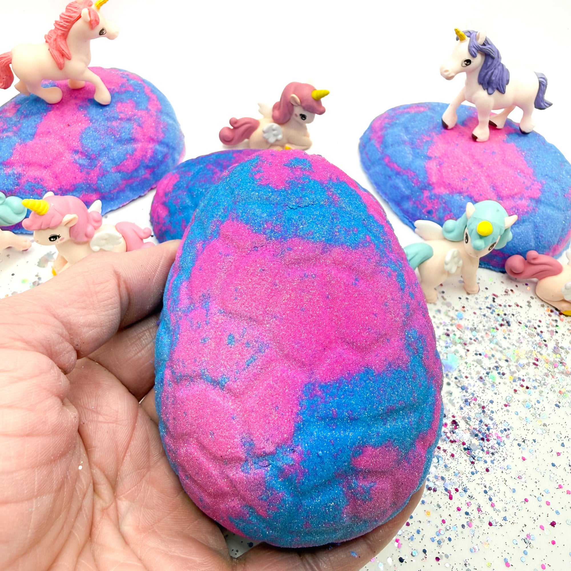 Blue and pink unicorn egg bath bomb held in a hand with unicorn figurines on a white background.