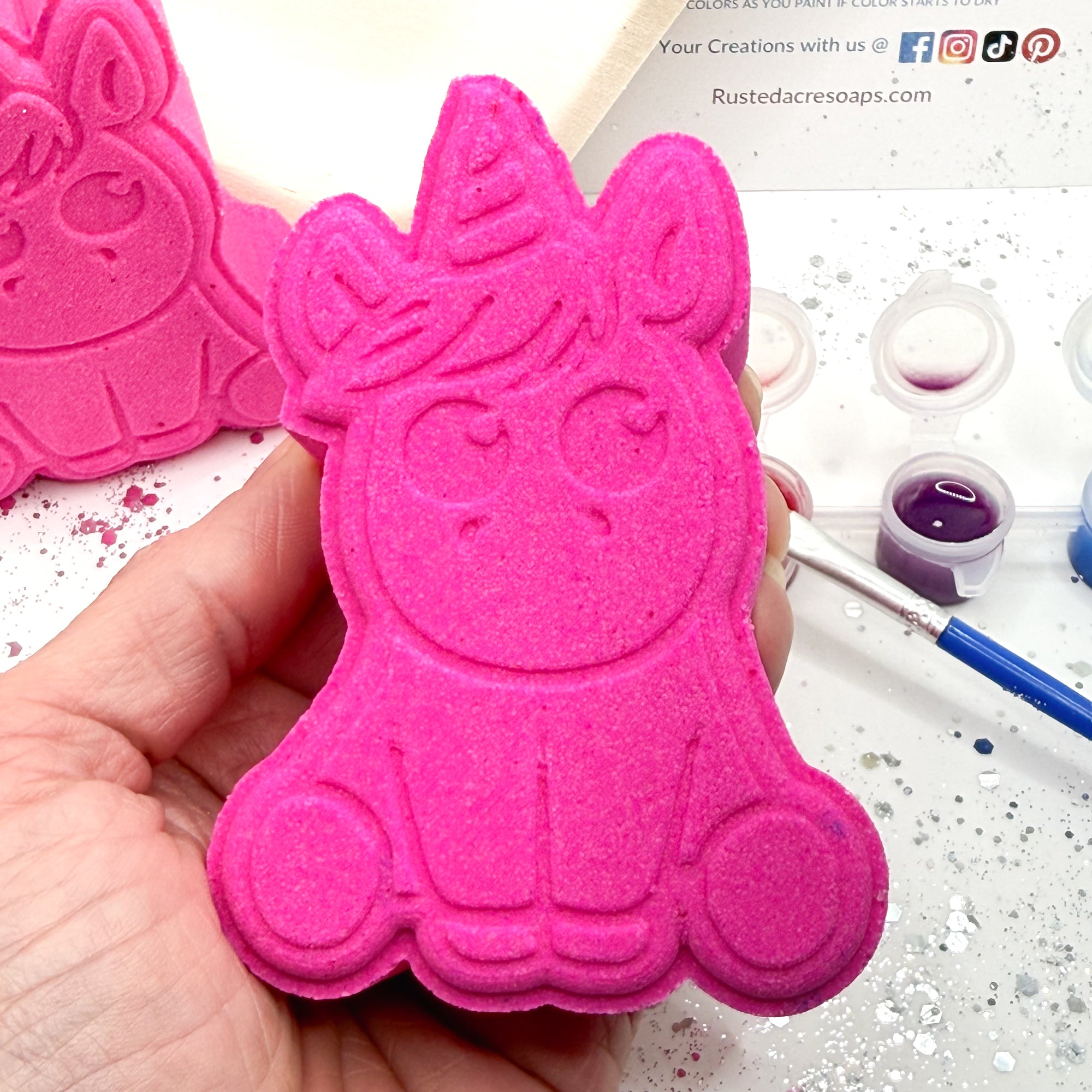 One pink paint your own unicorn shaped bath bomb held in hand with a paint brush, paint pots with color.