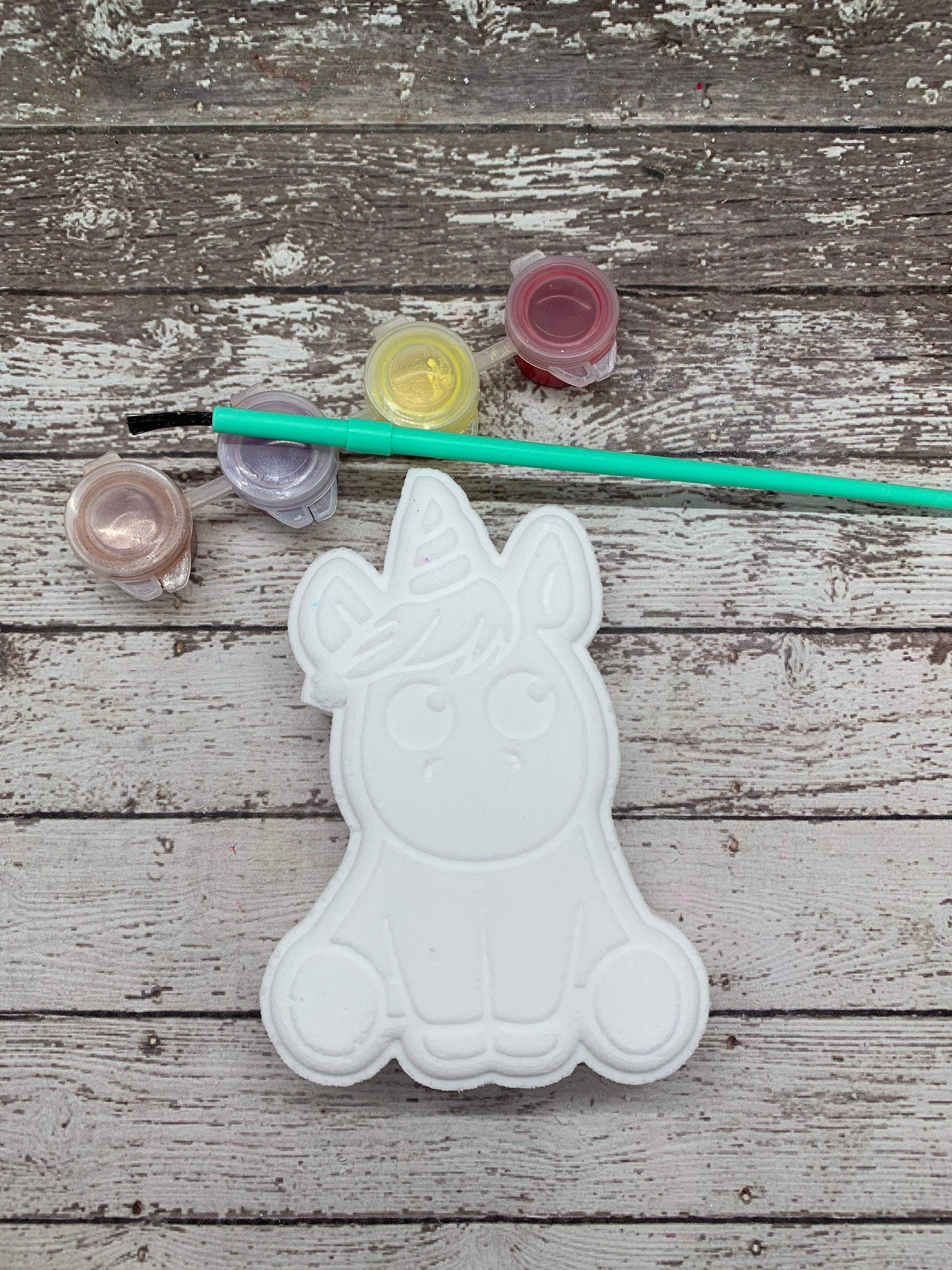 One white unicorn shaped shaped bath bomb displayed with a paint brush, paint pots with color.