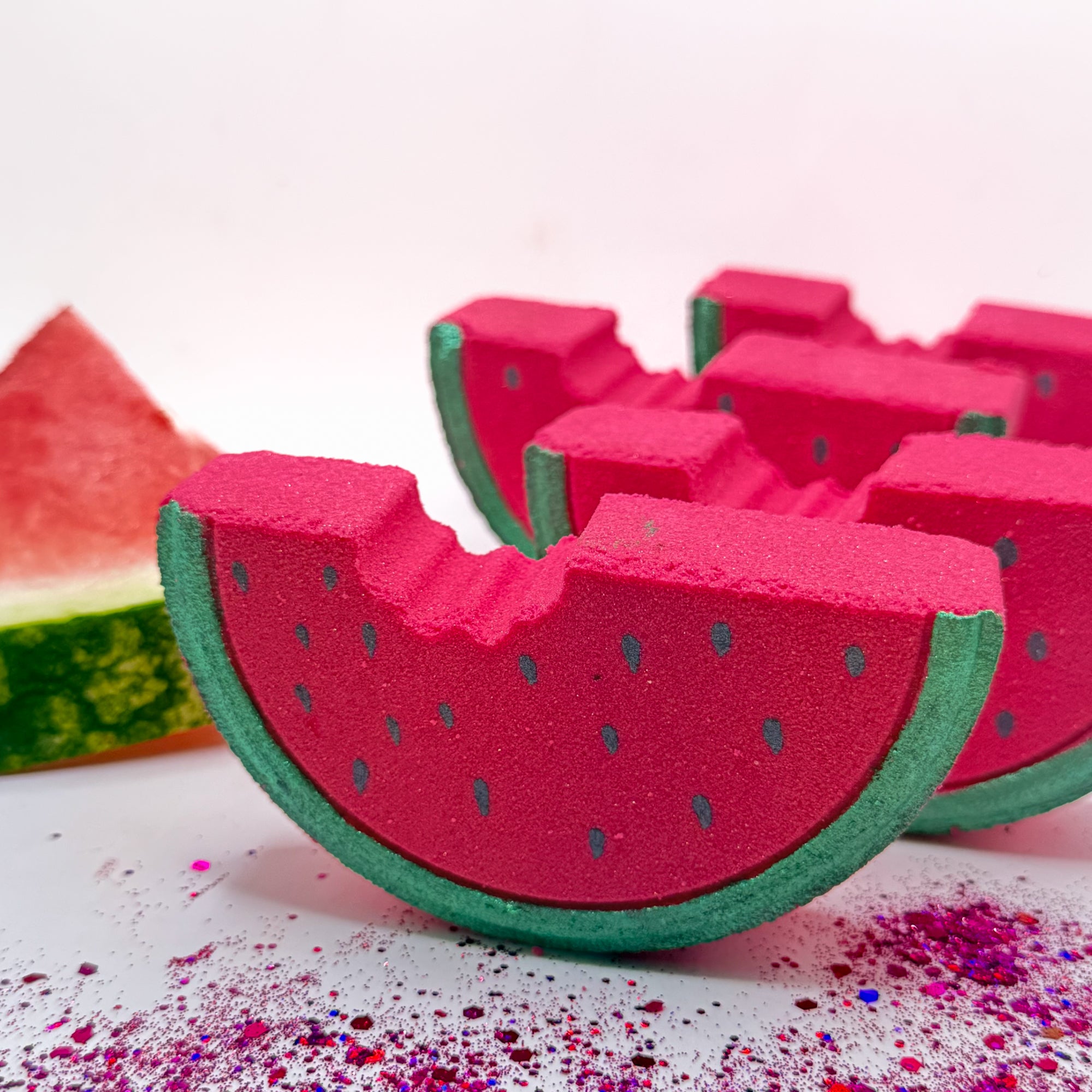Watermelon bath bombs in a staggered line on a white surface with colorful glitter and watermelon slice prop.