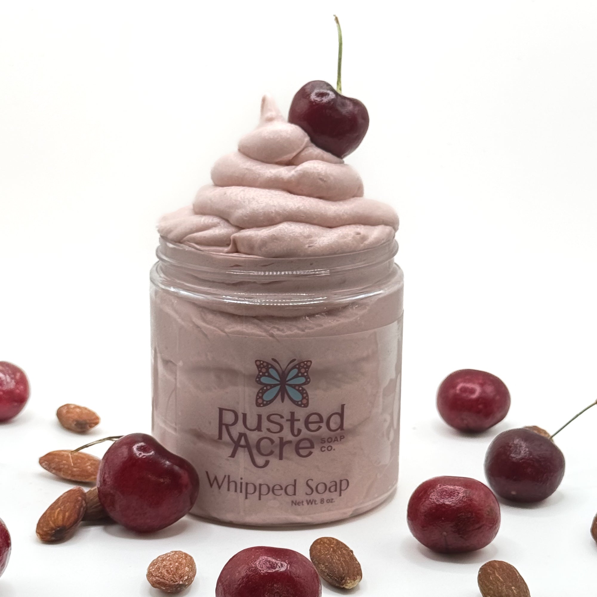 Jar of cherry almond whipped soap with a cherry on top, surrounded by cherries and almonds on a white background.