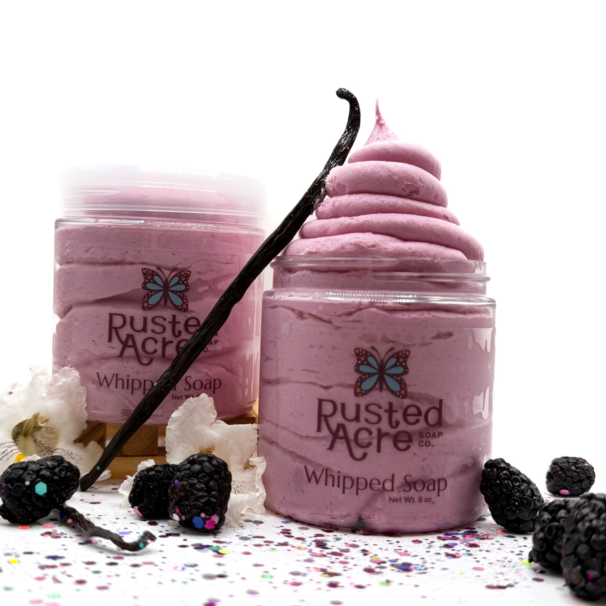 Two jars of Rusted Acre's pink raspberry vanilla whipped soap with blackberries and vanilla beans on a white background