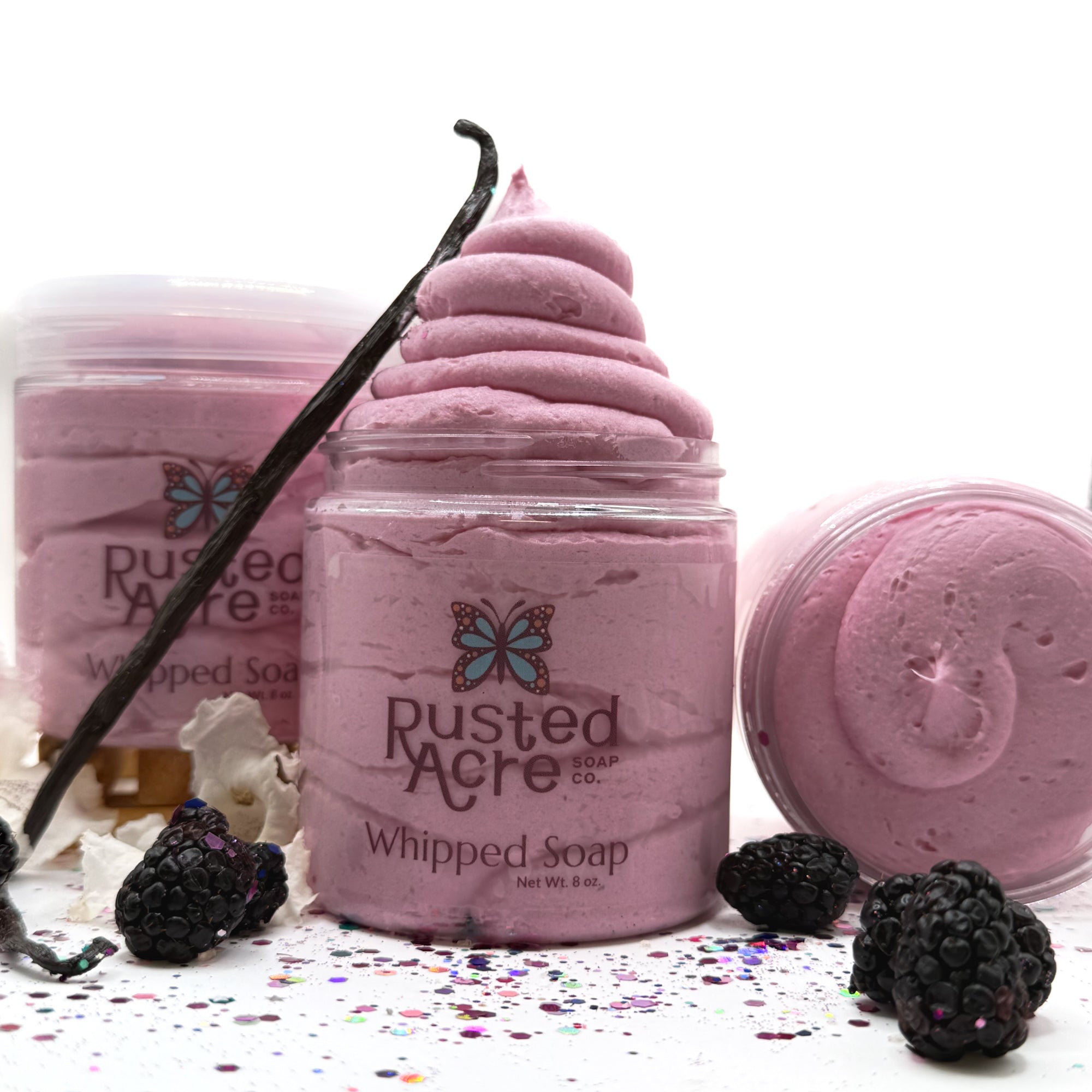 Three jars of pink raspberry vanilla whipped soap with vanilla bean, blackberries, and flowers on a white background