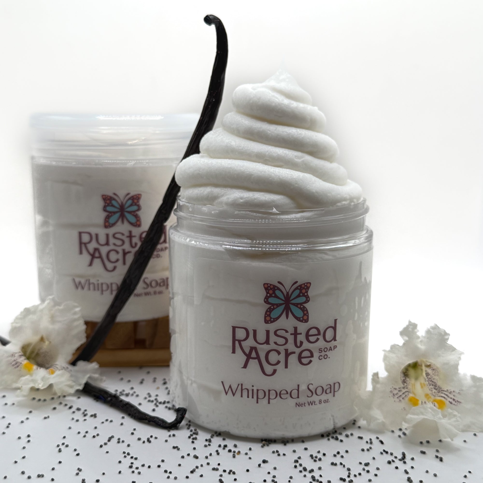 Rusted Acre vanilla whipped soap in a jar with a vanilla bean and flowers on a white background.