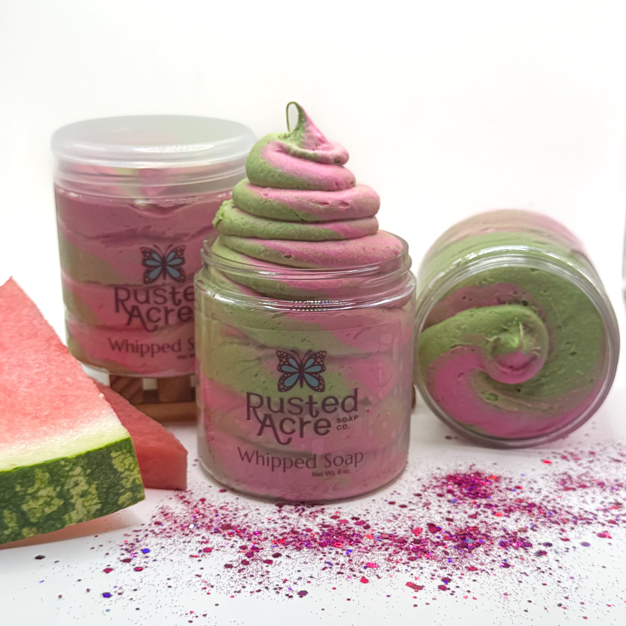 Three jars of Rusted Acre watermelon twist whipped soap with pink and green colored swirled soap, watermelon slice props, and sprinkles on a white background.