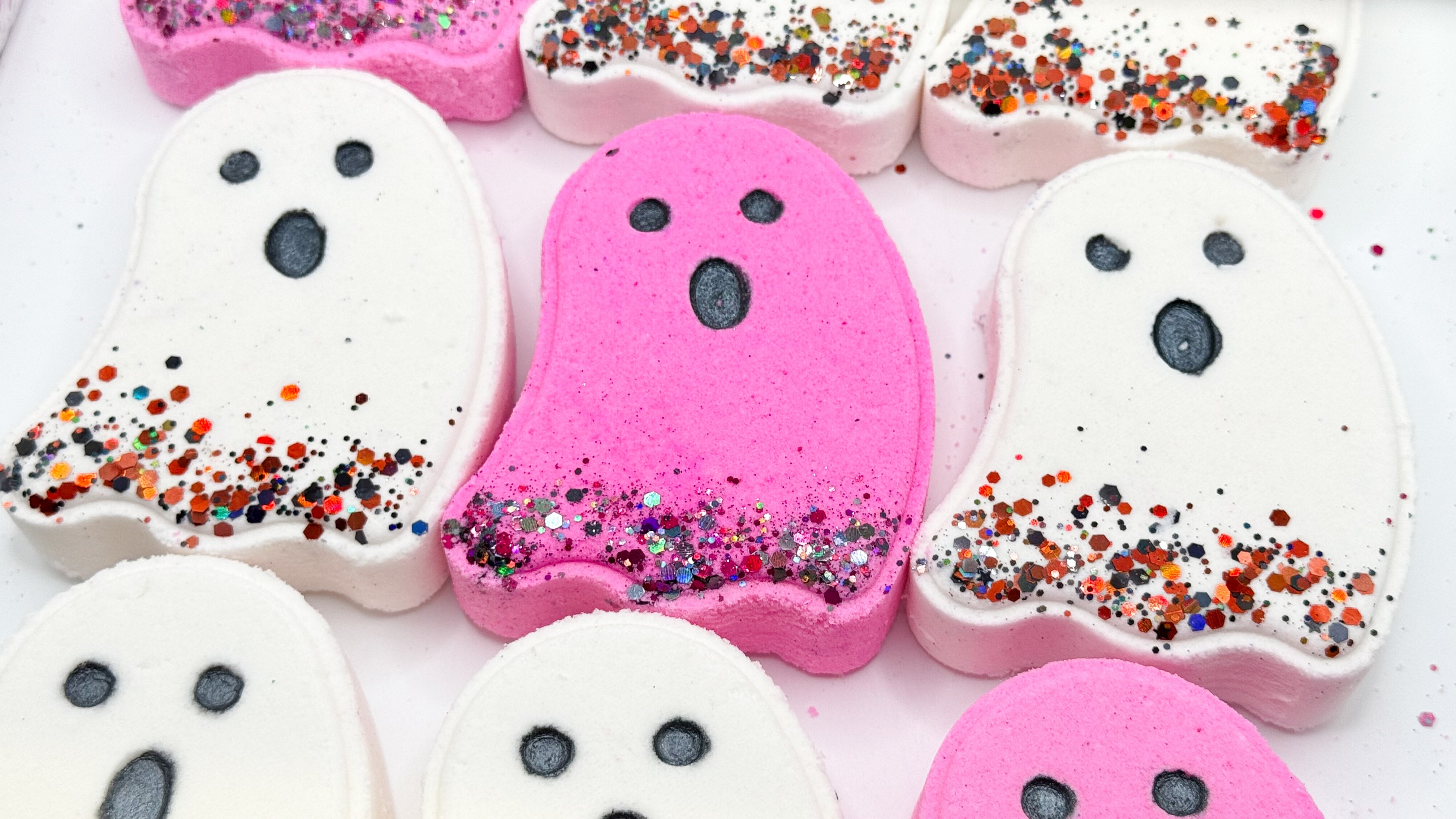 Pink and white ghost-shaped bath bombs decorated with black eyes and colorful orange, red, and silver glitter, arranged on a white background.