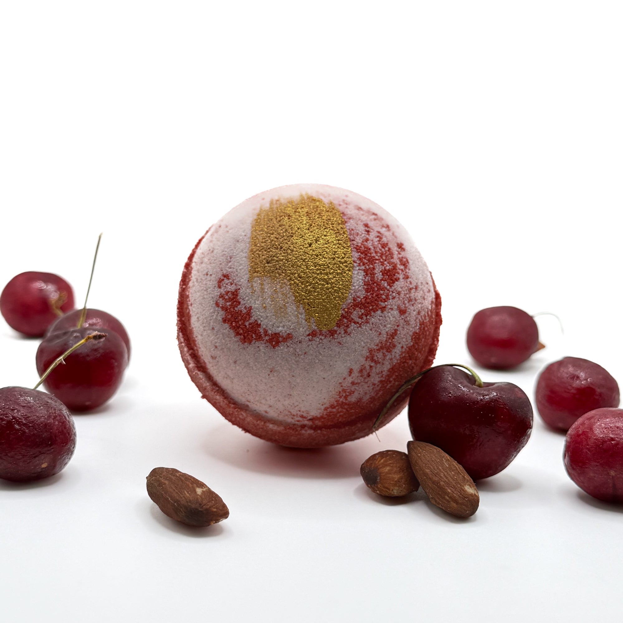 Handmade cherry almond bath bomb with gold mica paint surrounded by red cherries and almonds on a white background.
