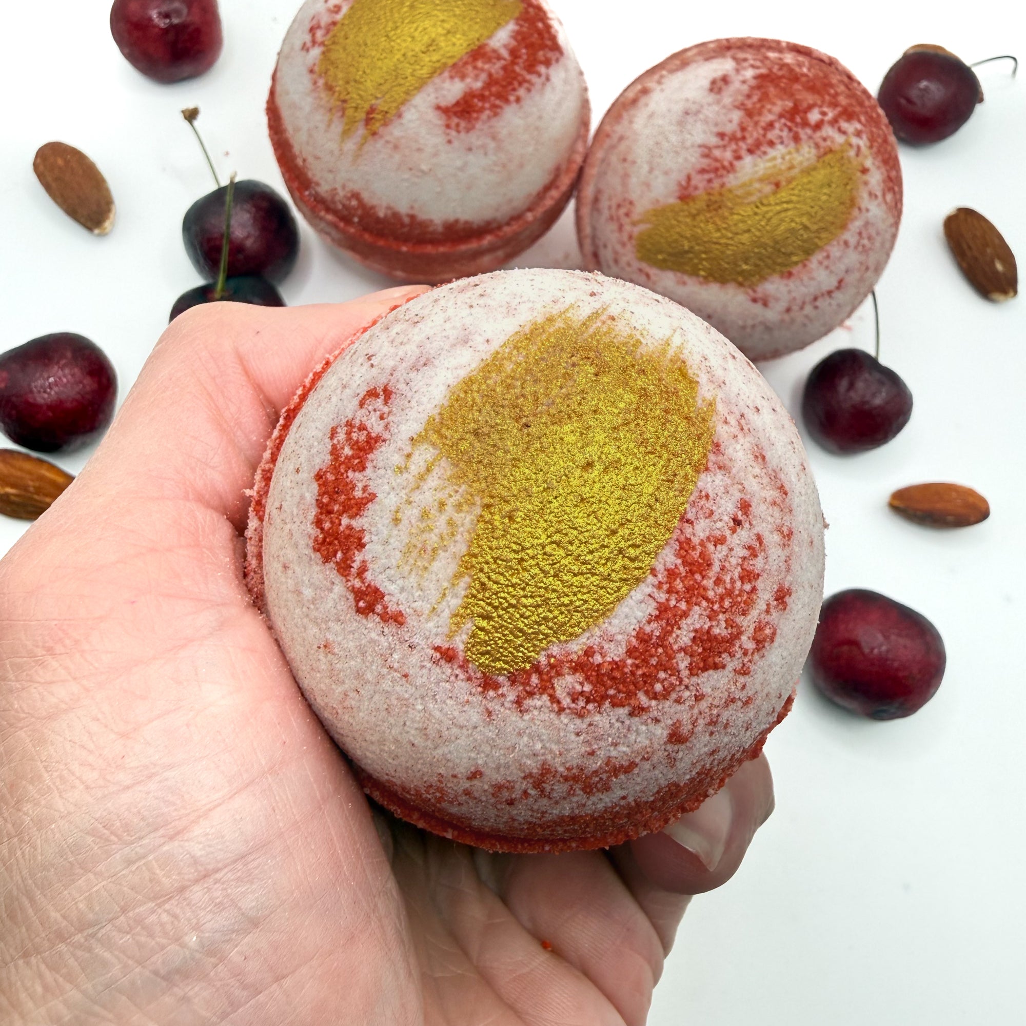 3 handmade cherry almond bath bombs swirled red and white with gold mica paint in a hand by Rusted Acre Soap Co.
