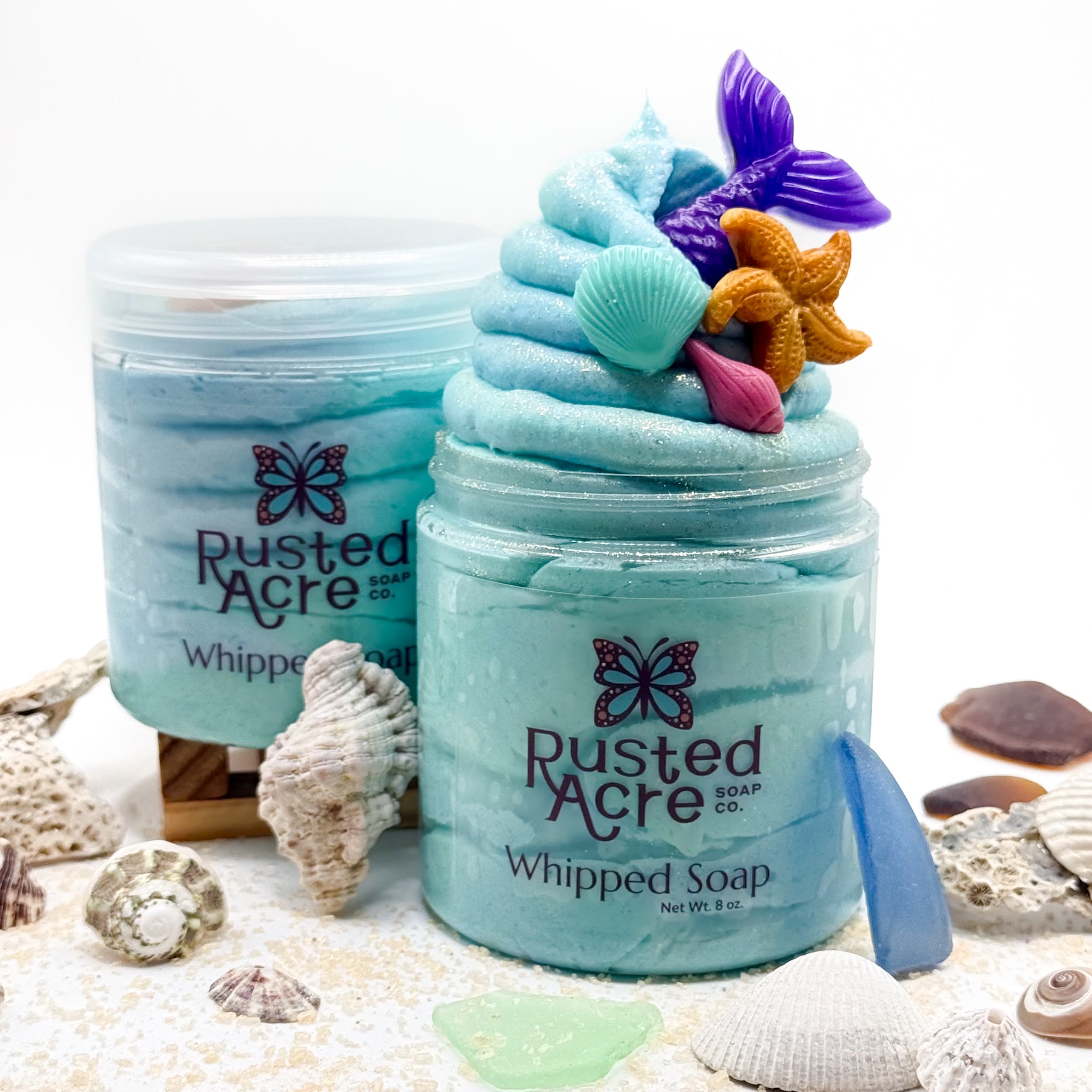 Two jars of blue mermaid kiss whipped soap by Rusted Acre surrounded by seashells. 