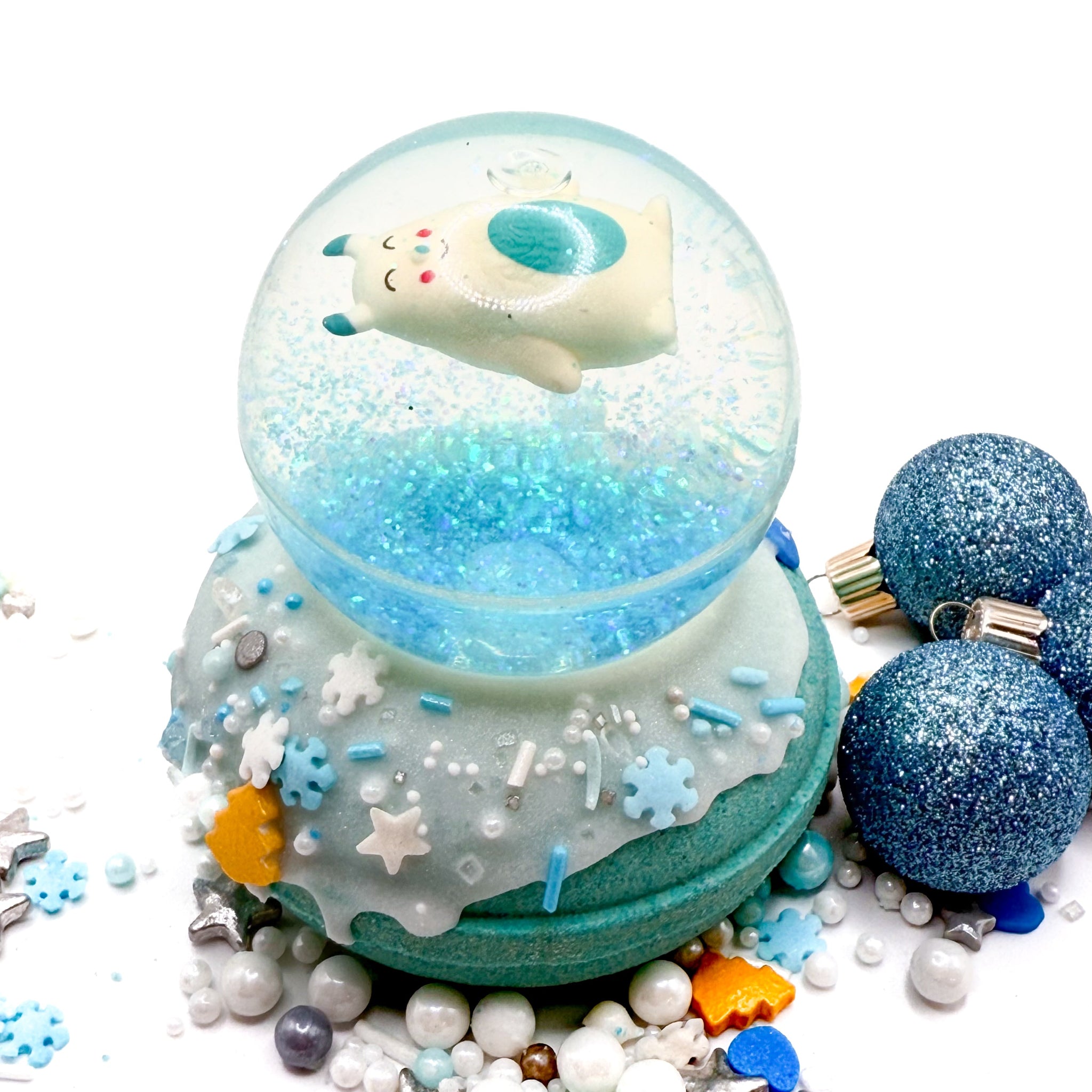 Snow globe bath bomb topped with an Abominable Snowman bouncy ball, Blue balls and silver sprinkles in the background. Handmade by Rusted Acre Soap Co.