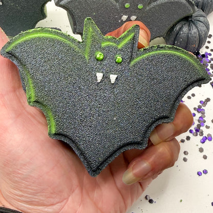 A hand holding a bat shaped Halloween bath bomb, black with bright green edging, green eyes, and white fangs, with glitter decorations in the background.
