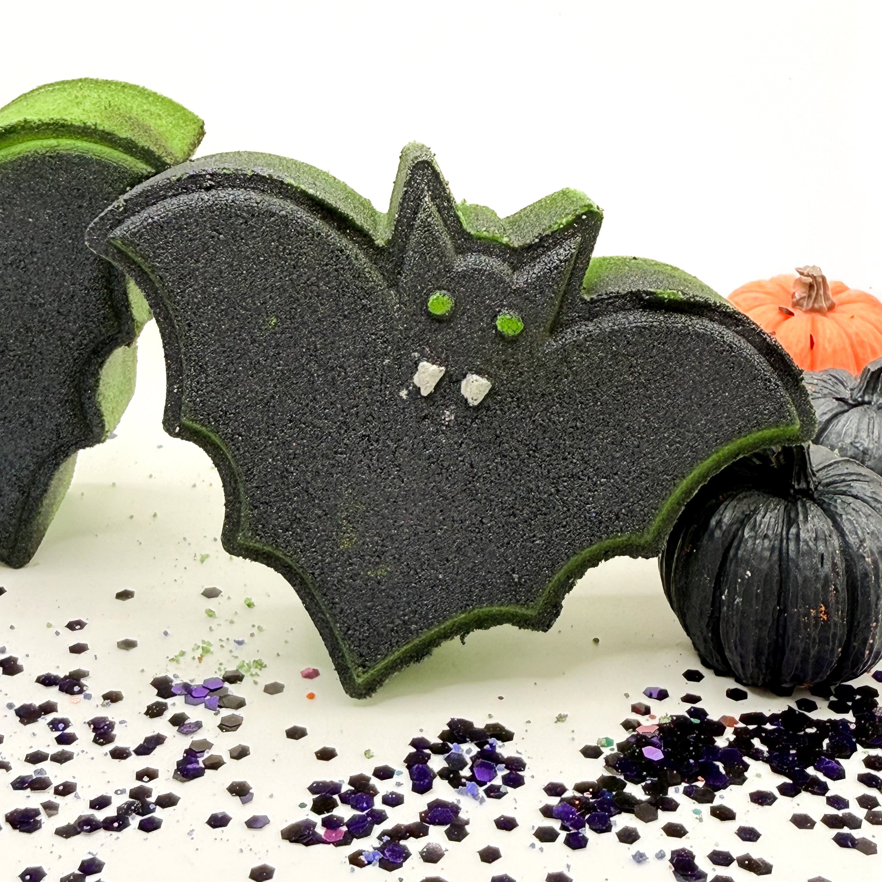 A Halloween bat bath bomb with a black glittery body, green edges, and small white teeth details, displayed alongside other bath bombs and decorative black pumpkins on a white surface with purple glitter.