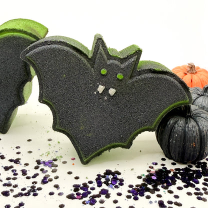 A Halloween bat bath bomb with a black glittery body, green edges, and small white teeth details, displayed alongside other bath bombs and decorative black pumpkins on a white surface with purple glitter.