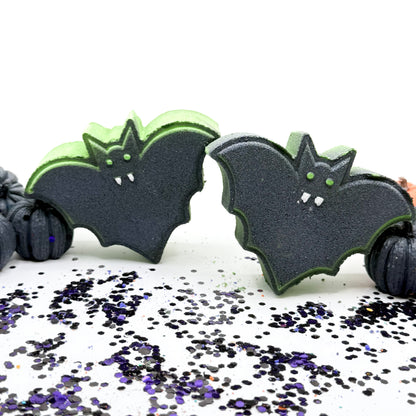 Two bat bath bombs with a black body and bright lime green edges, decorated with small white fangs and green eyes, displayed on a white surface scattered with black and purple glitter.