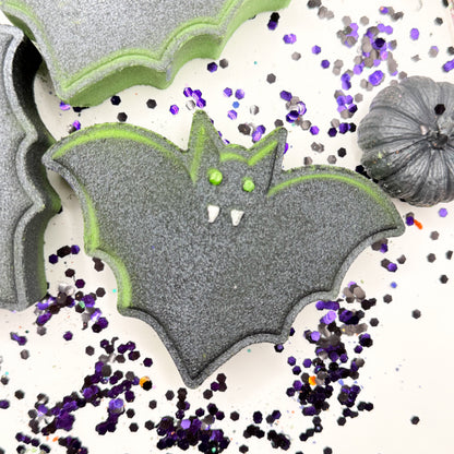 A bat bath bomb with lime green edges, purple eyes, and white fangs, shown on a white background with scattered purple and black glitter and a small gray decorative pumpkin.