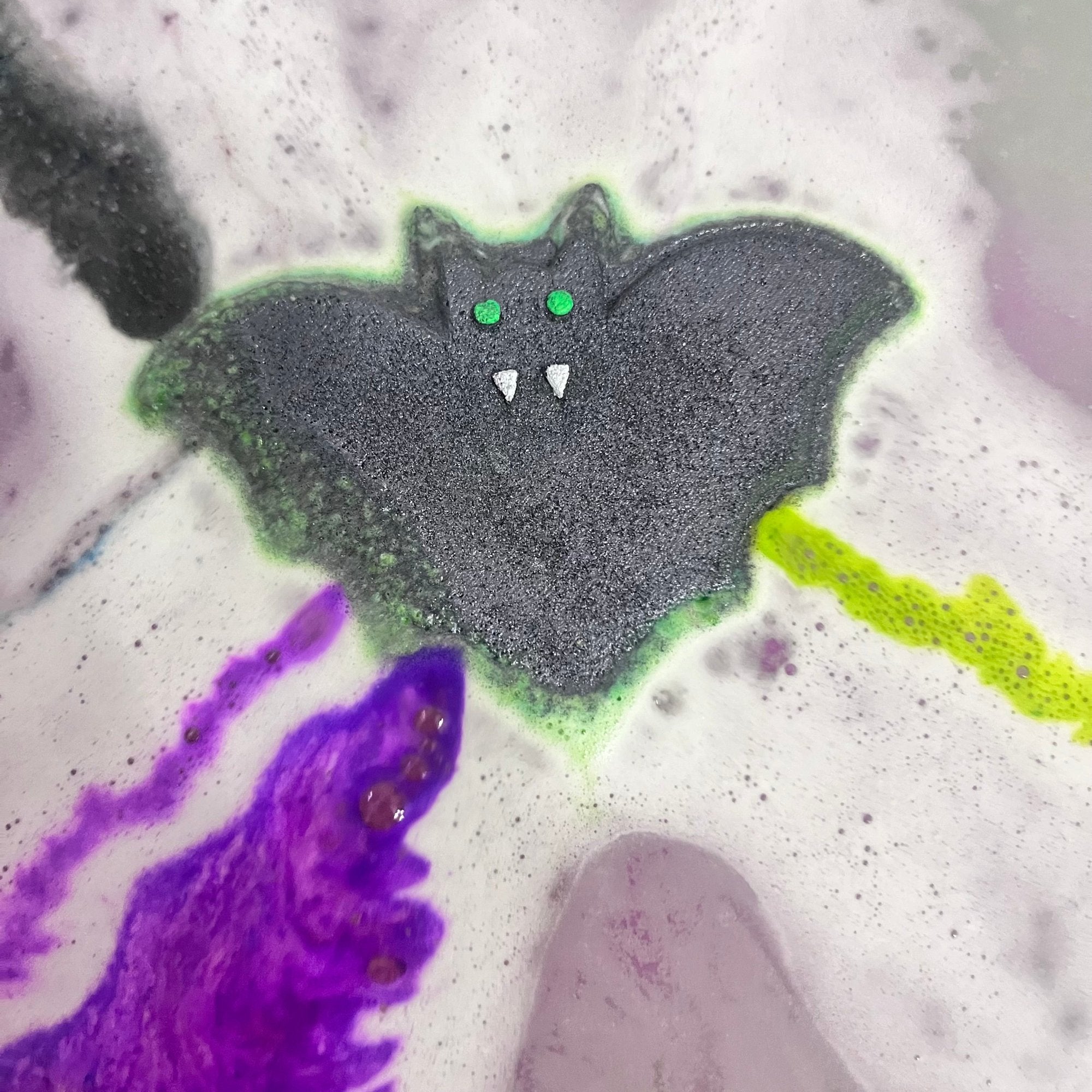 A green bat shaped bath bomb airbrushed black with hand-painted green eyes and white fangs in water. Purple and green streaks of fizz and foam coming out of the sides!
