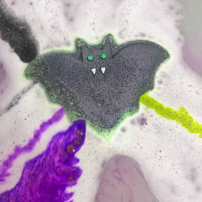 A green bat shaped bath bomb airbrushed black with hand-painted green eyes and white fangs in water. Purple and green streaks of fizz and foam coming out of the sides!