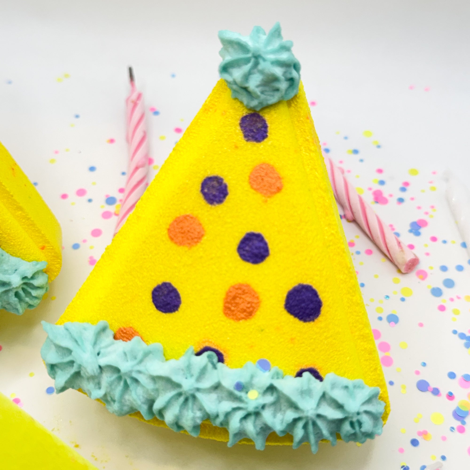 One yellow birthday hat shaped bath bomb with hand painted polka dots laying on a white background with colorful glitter dots. 