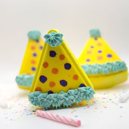 Yellow birthday hat shaped bath bombs with hand painted polka dots standing up on a white background with colorful glitter. 
