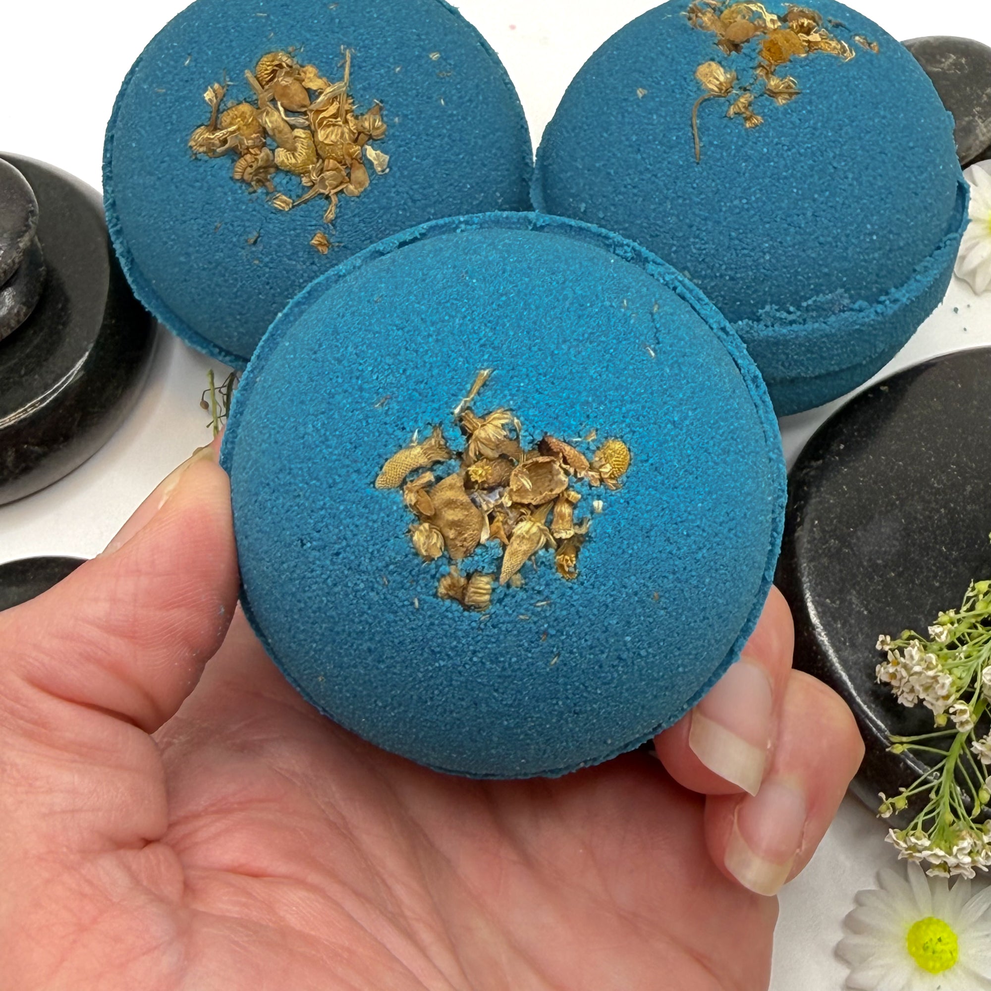 Three blue tranquility bath bombs, one is held in a hand to show the dried flower details. 