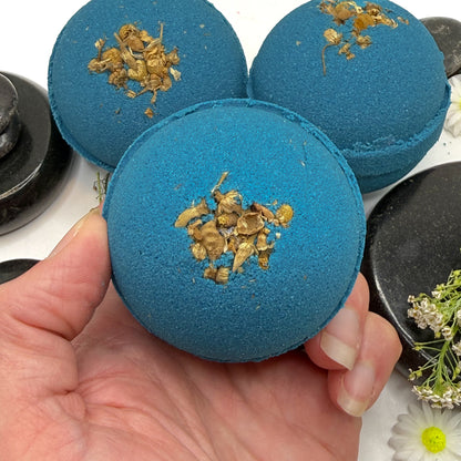 Three blue tranquility bath bombs, one is held in a hand to show the dried flower details. 