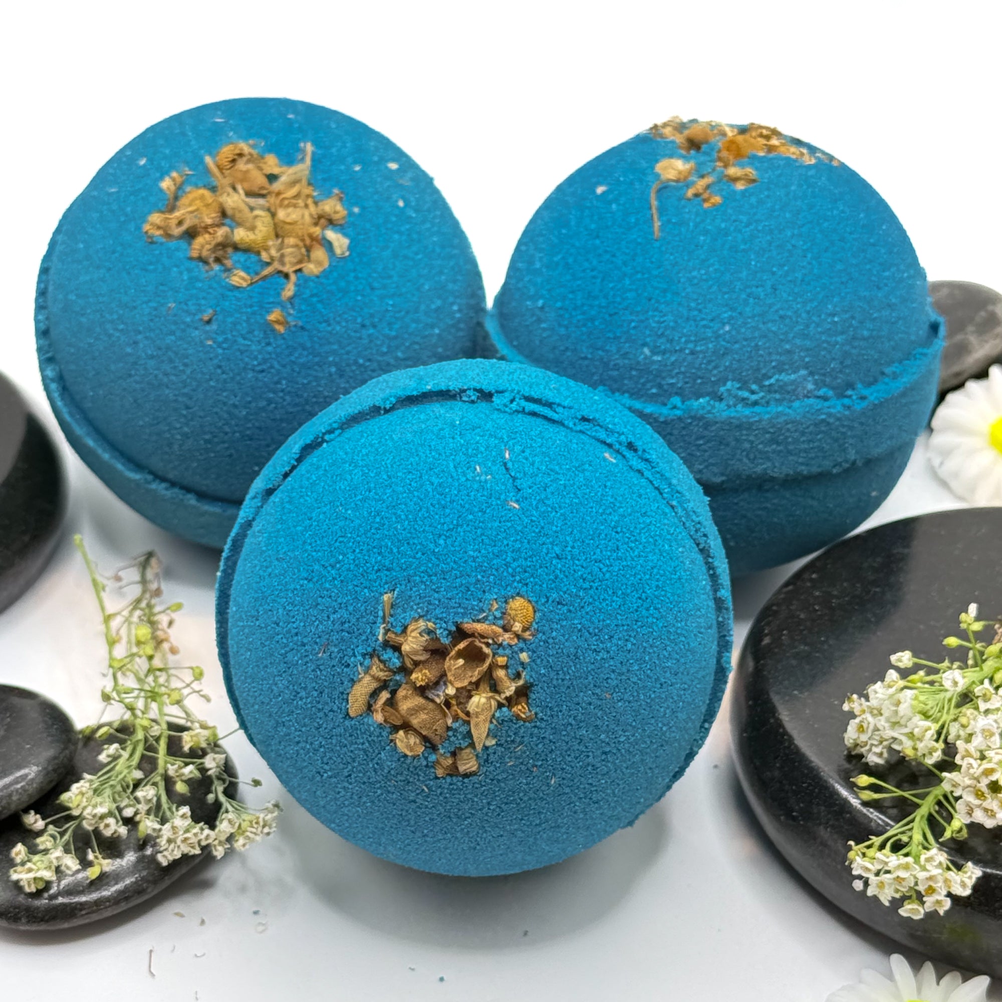 Three blue tranquility bath bombs with dried flower details on a white background with spa type props. 