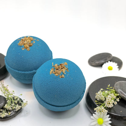 Twoblue tranquility bath bombs with dried flower details on a white background with spa type props. 