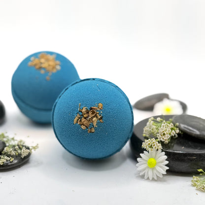 Two blue tranquility bath bombs with dried flower details on a white background with spa type props. 