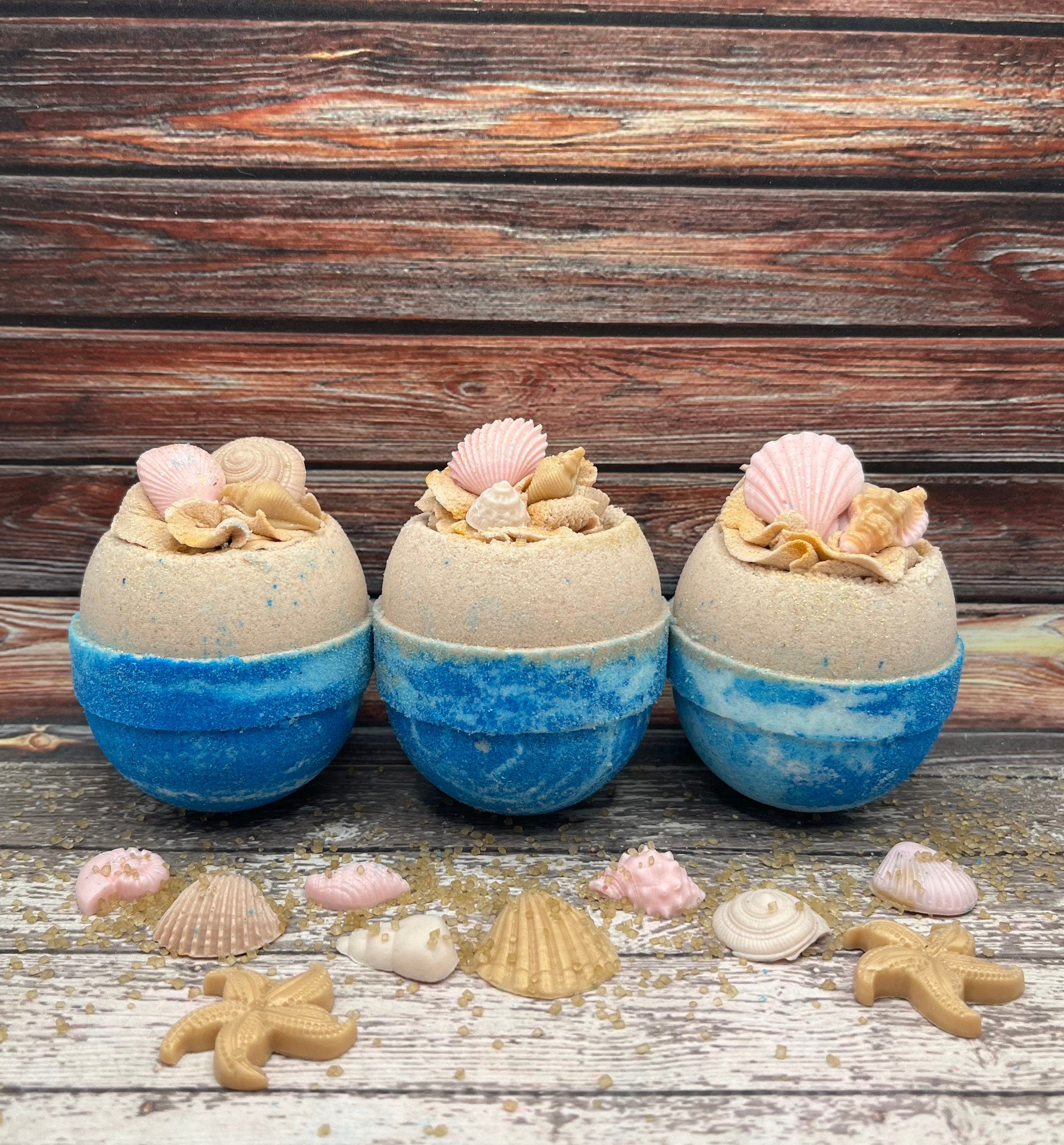 Three beach themed round bath bombs. Light-blues swirled bottom, tan upper half topped with piped bubble frosting embedded with with sea shell shaped soaps