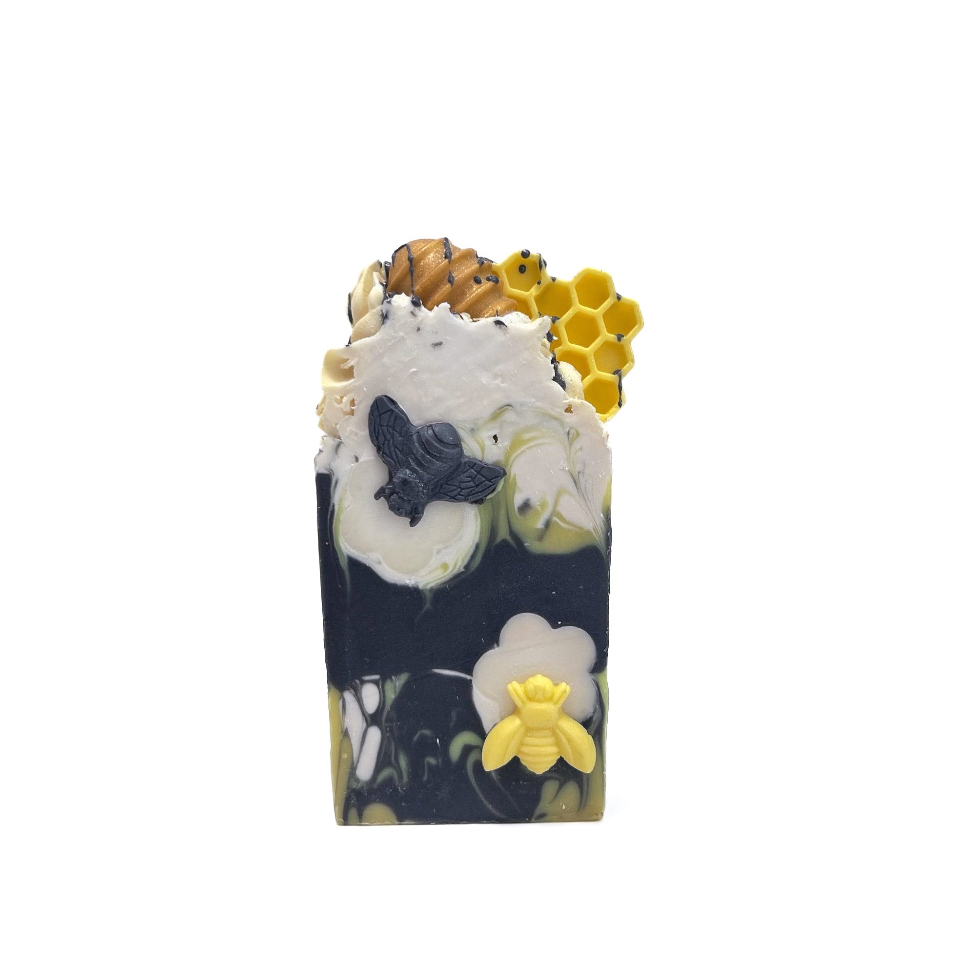 A bar of Rusted Acre Soap Company's Bee Happy handmade soap. Swirled charcoal black, white, and yellow. A yellow and a black bee soap embed on front. Topped with a  honey comb and dipper soap embed. 