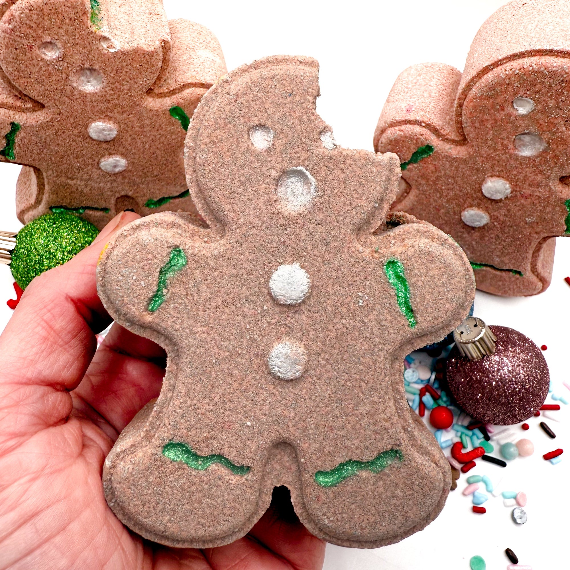 Decorative hand painted gingerbread man bath bomb held by a hand with Christmas decorations in the background.