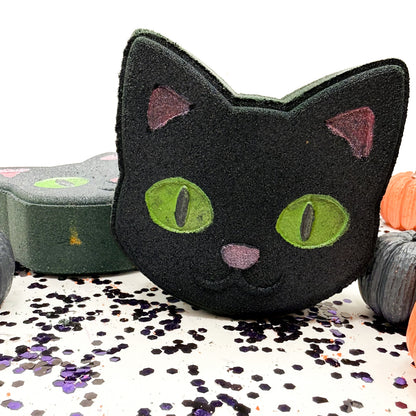 A black cat-shaped bath bomb with green eyes and pink nose, displayed on a white surface scattered with purple and black glitter, with other Halloween themed soaps in the background.