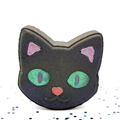 Rusted Acre Soap Co.’s handcrafted Black Cat Bath Bomb standing upright, shaped like a cat’s face with bright green eyes, pink ears, and a red nose, shown against a white background with scattered blue and purple confetti.