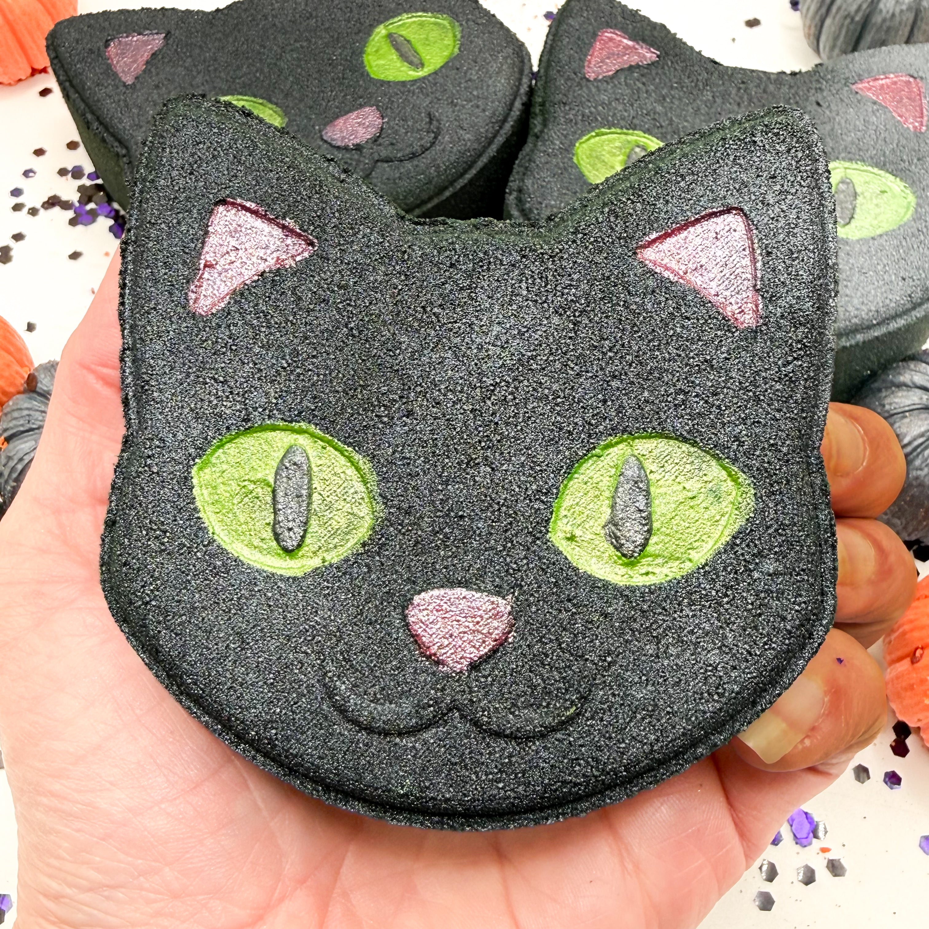 A hand holding a black cat shaped bath bomb with glittery green eyes, pink nose, and pink ears, with more similar cat bath bombs in the background.