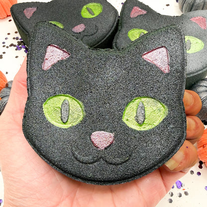 A hand holding a black cat shaped bath bomb with glittery green eyes, pink nose, and pink ears, with more similar cat bath bombs in the background.