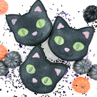 Three black cat bath bombs with green eyes, pink noses, and pink ears, arranged on a white background with small pumpkins and purple sprinkles.